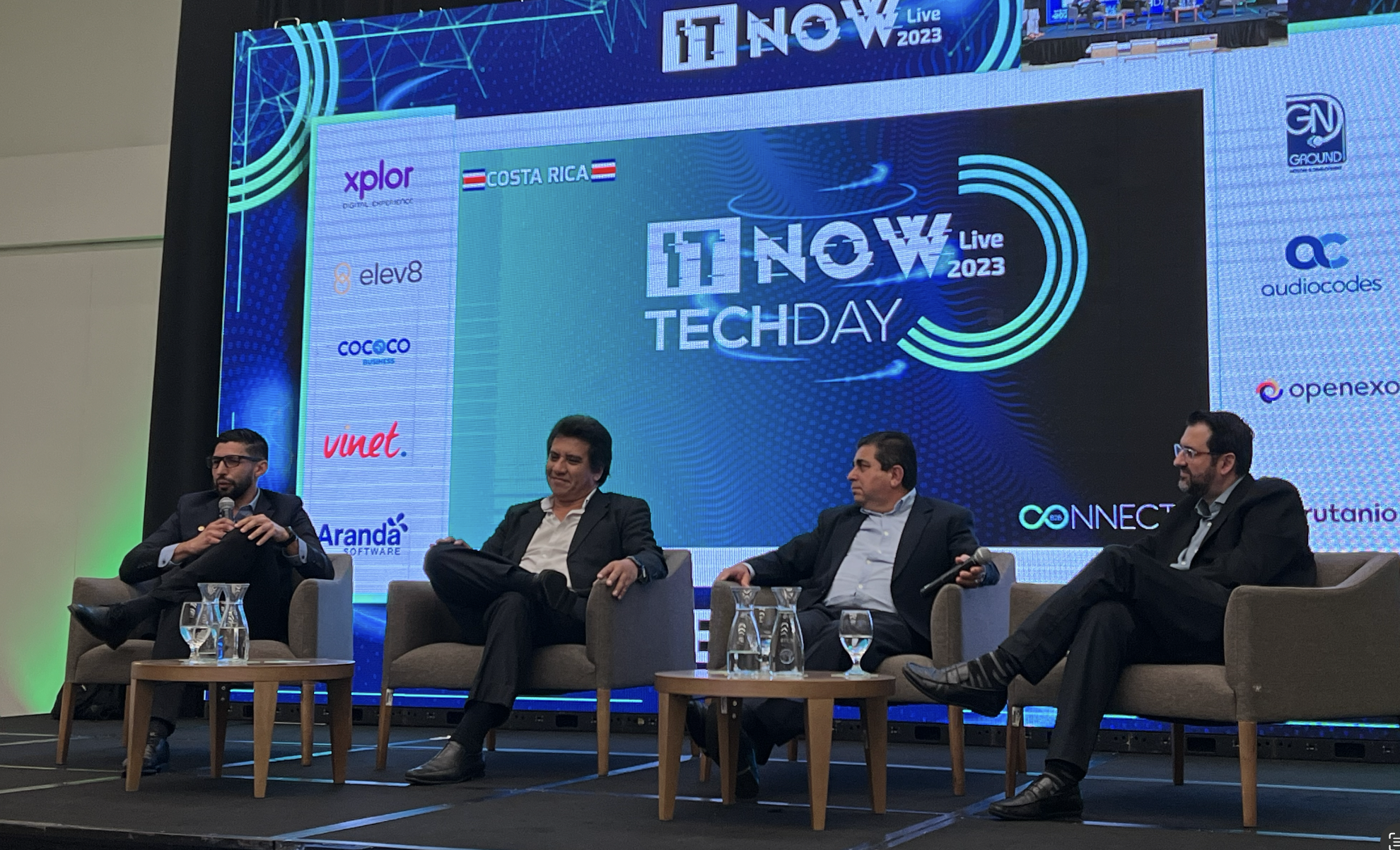 Fotografía plano general de los panelistas con la imagen de fondo del logo del Tech Day 