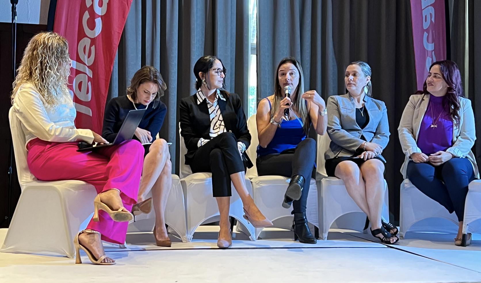 Panelistas sentadas en sillas blancas, moderadora más cinco panelistas, en el medio con el micrófono Rebeca Esquivel